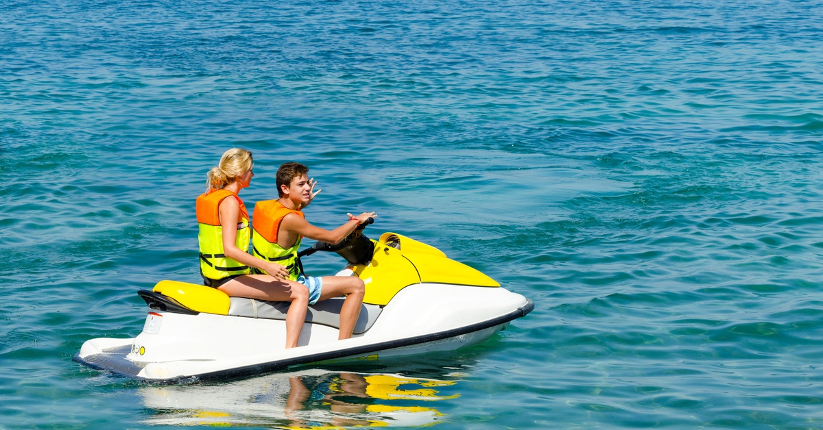 A man and a woman in yellow and orange life jackets sit on a yellow and white jet ski on calm water.
